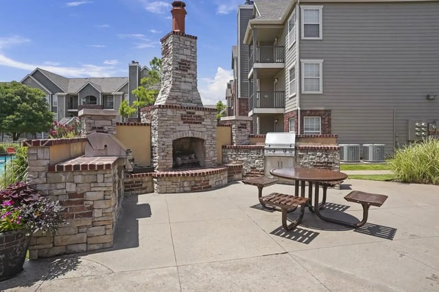 Outdoor community grilling area with stone fireplace and table, surrounded by apartment buildings.