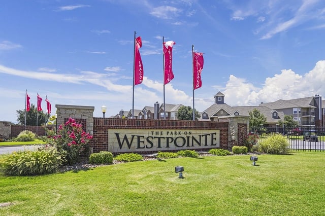 Entrance sign for The Park on West Pointe with brick pillars, landscaping, and red flag banners.