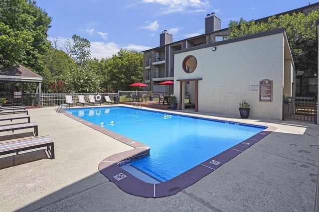 Outdoor apartment community pool with lounge chairs, red umbrellas, and a small clubhouse.