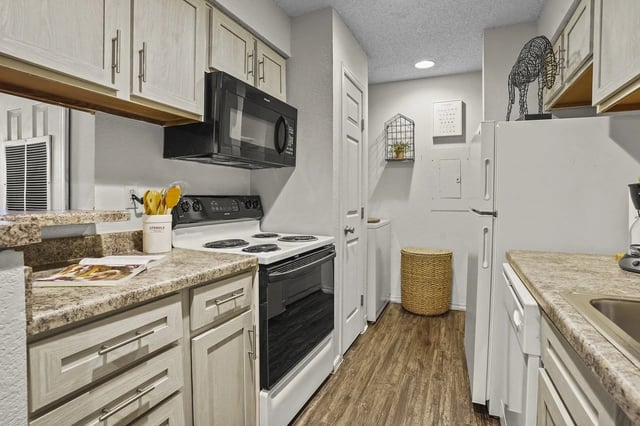 Galley kitchen with white cabinets, beige countertops, stove, microwave, and refrigerator in an apartment.