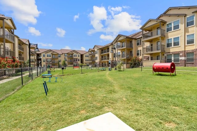 Apartment complex courtyard with a fenced-in dog park featuring agility equipment like tunnels and hoops.