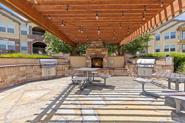 Outdoor grilling area with built-in grills, fireplace, and picnic tables under a wooden pergola.