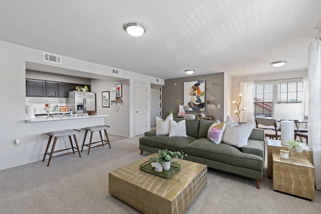 Open concept living and kitchen area with green sectional sofa, gold coffee table, and kitchen island with bar stools.