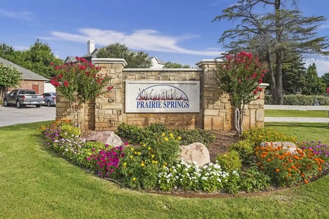 Stone entrance sign with Prairie Springs branding, flanked by colorful flower beds and trees.