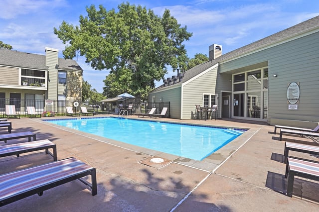 Swimming pool area with lounge chairs and tables next to apartment buildings.