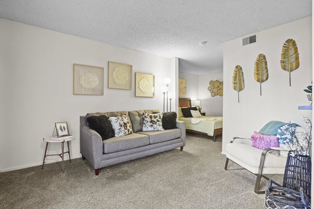 A staged living room and bedroom area with a gray sofa, white armchair, and a bed.
