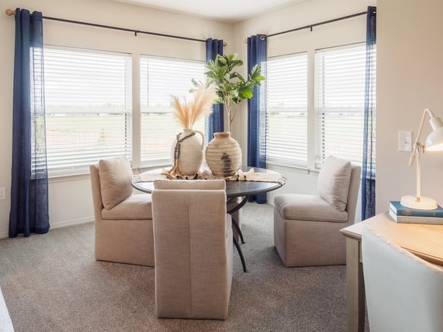 Sunlit dining area with a round table and four beige upholstered chairs by large windows with blue curtains.