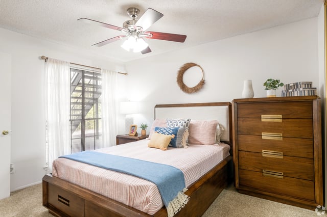 Queen bed with white bedding and pink stripes, a blue throw blanket, and decorative pillows. A wooden nightstand with a lamp and a dresser are also visible.