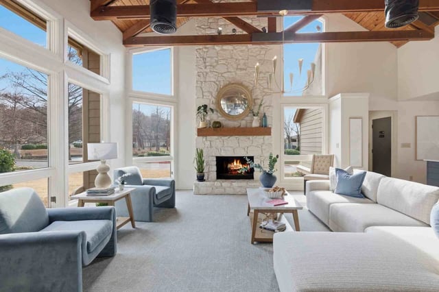 Bright living room with a stone fireplace, vaulted wooden beams, and large windows.