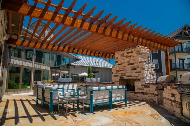 Outdoor community lounge with a stone fireplace, grills, and cushioned seating under a wooden pergola.