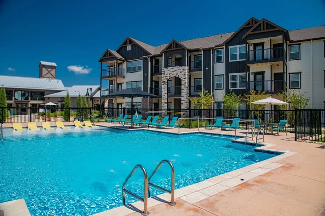 Outdoor community pool with blue water, lounge chairs, and an apartment building in the background.