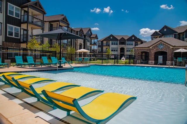 Outdoor apartment community pool with blue water, yellow lounge chairs, and surrounding buildings.