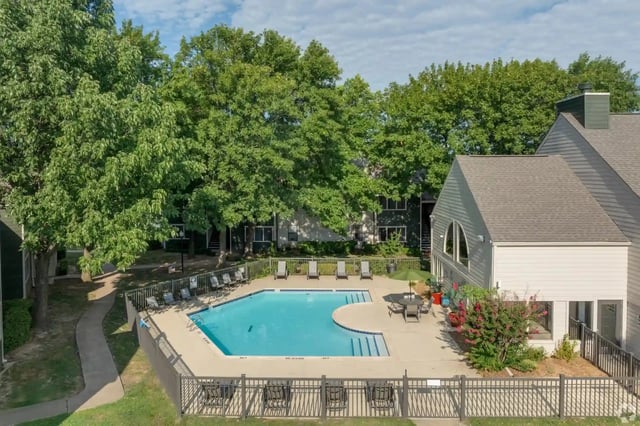 Outdoor pool area with lounge chairs around a blue pool, shaded by large trees.