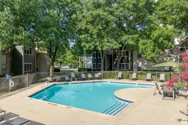 Outdoor apartment pool with lounge chairs under leafy trees.