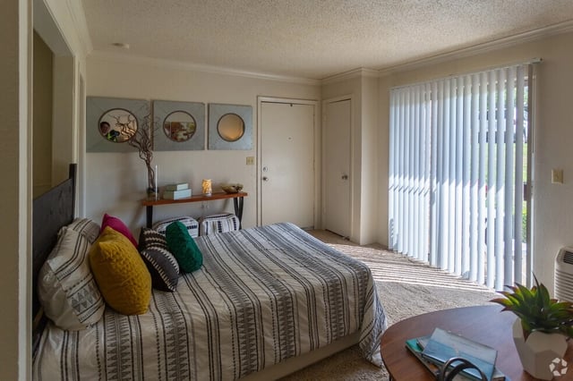 Bedroom with a queen-size bed, decorative pillows, and vertical blinds covering a sliding glass door.