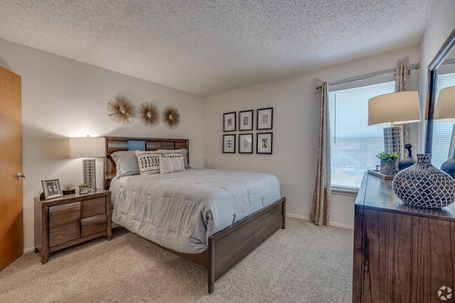 Queen bed with gray comforter and decorative pillows, nightstand, dresser, and window in a bedroom.