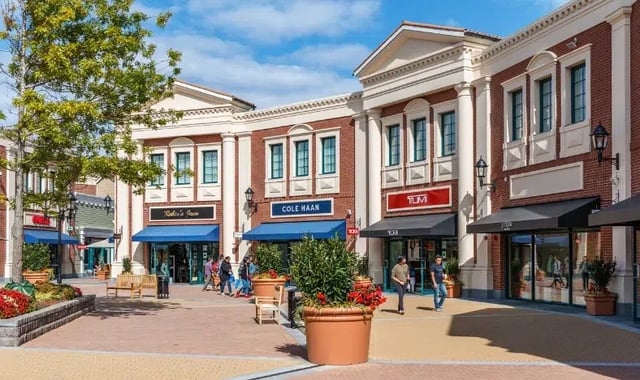 Outdoor plaza with brick storefronts, planters, awnings, and pedestrians under a blue sky.
