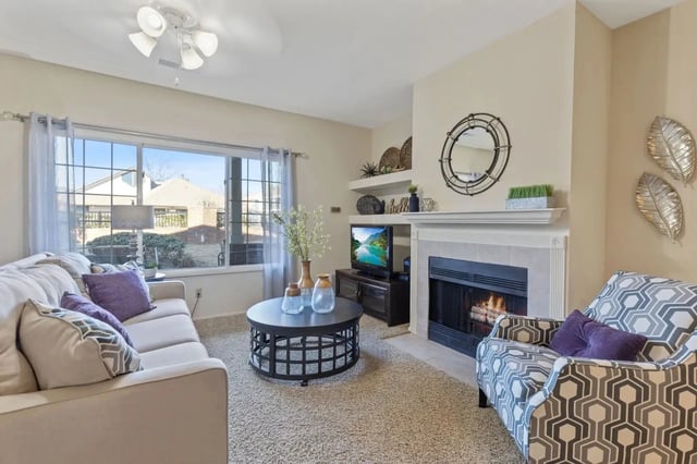 Living room with beige sofa, patterned chair, coffee table, fireplace, and large window with sheer curtains.