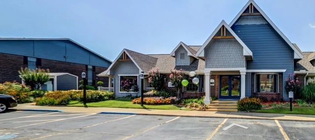 Exterior view of the apartment building entrance with landscaping and parking lot.