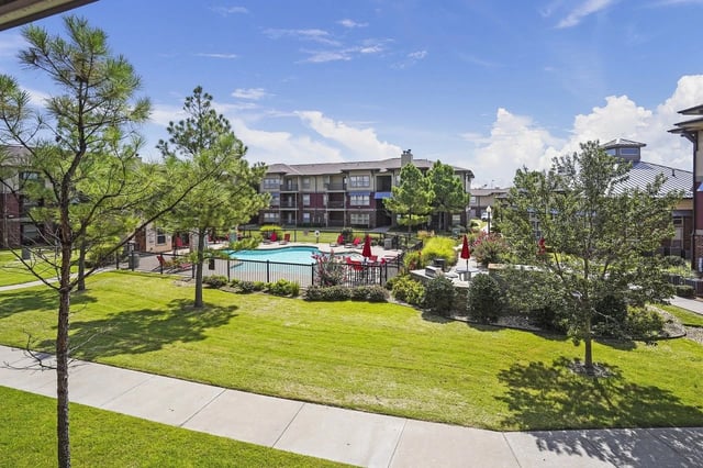 Outdoor apartment community pool area with lounge chairs and red umbrellas, surrounded by trees.