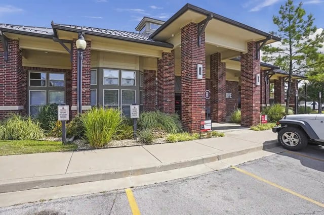 Exterior view of a brick apartment building with columns, landscaping, and a sidewalk in front.