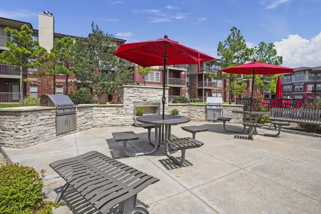 Outdoor communal courtyard with grills, tables, benches, and red umbrellas near apartments.