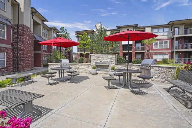 Outdoor apartment courtyard with red umbrellas, tables, built-in grills, and stone accents.