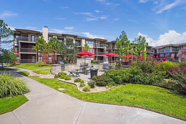 Outdoor grilling area with picnic tables and red umbrellas in front of apartment buildings.