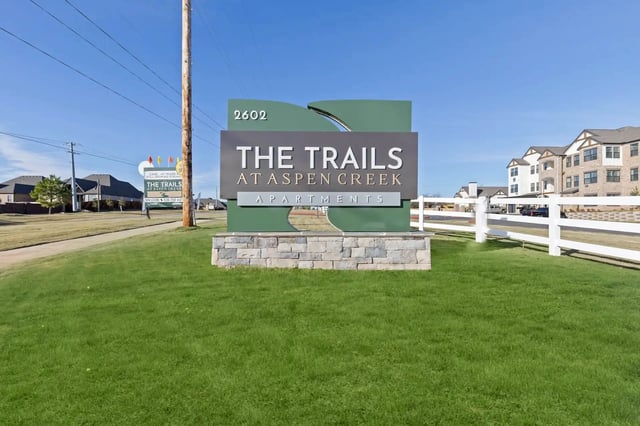 Front entrance sign for The Trails at Aspen Creek Apartments on a green lawn with a white fence.