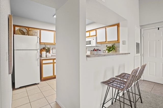 Kitchen area with white refrigerator, white cabinets, and a breakfast bar with two woven chairs.