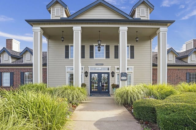 Front entrance of a beige apartment building with tall columns and landscaping.