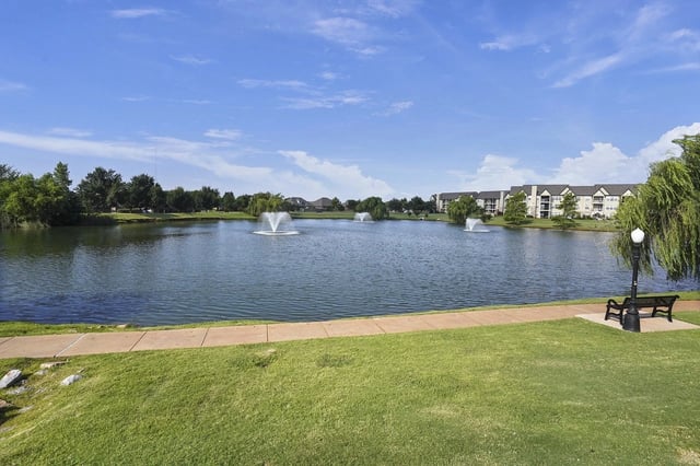 Pond with fountains, grassy park, and a bench along a walkway with apartment buildings in the distance.