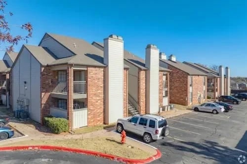 Exterior view of a brick apartment complex with multiple entrances, a parking lot, and clear blue sky.