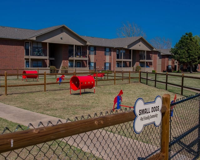 Exterior view of brick apartment buildings with a fenced dog park and playground in the lawn.