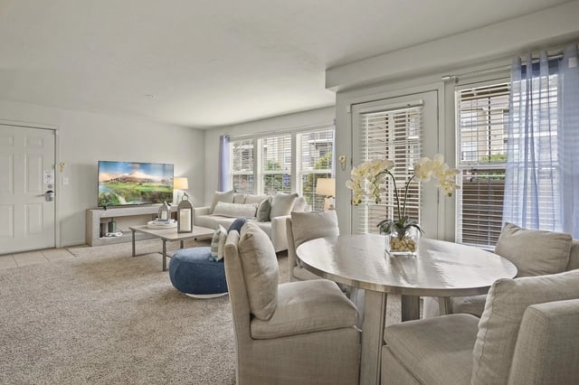 Bright apartment living room with beige couch, round dining table, TV, and large windows with blinds.