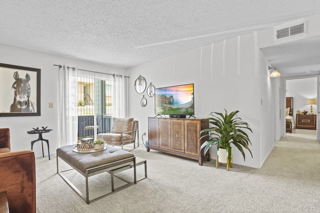 Apartment living room with TV on a wooden stand, seating, plant, and sliding door to balcony.