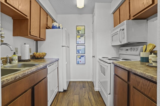 Compact apartment kitchen with white appliances, wooden cabinets, and a double sink.