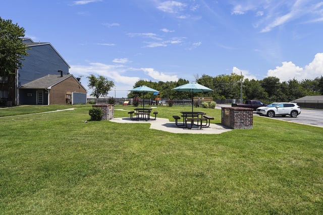 Outdoor picnic area with two tables, umbrellas, and grills next to apartment buildings and a parking lot.