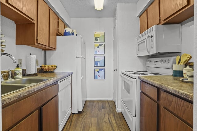 Compact apartment kitchen with white appliances, wooden cabinets, and a double sink.