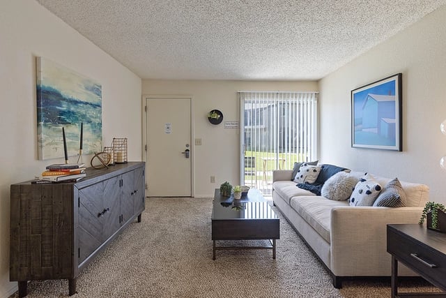 Living room in an apartment with a beige sofa, coffee table, wall art, and a sliding glass door to the balcony.