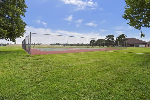 Fenced outdoor court with grassy foreground and a clubhouse in the background.