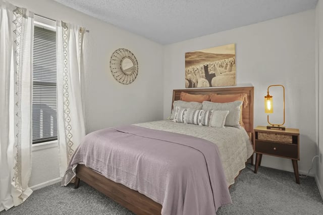 Bedroom with a wooden bed, lavender blanket, pillows, nightstand lamp, and window with curtains.