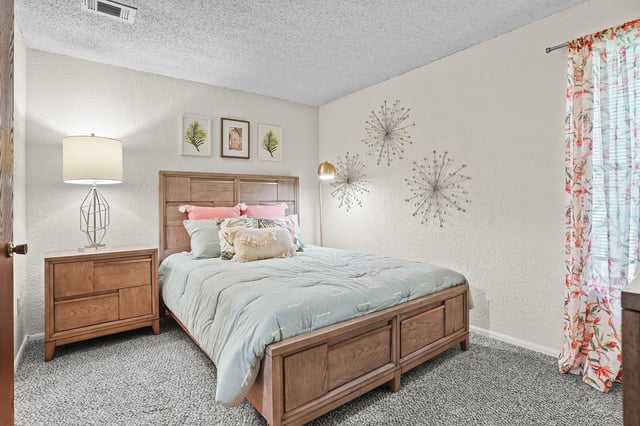 A cozy bedroom featuring a queen-size bed with light blue bedding, plush pillows, and a wooden headboard. A nightstand with a modern lamp sits to the left, and decorative starbursts adorn the wall.