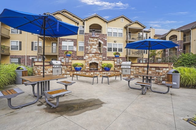 Outdoor seating area with picnic tables, umbrellas, grills, and a stone fireplace.