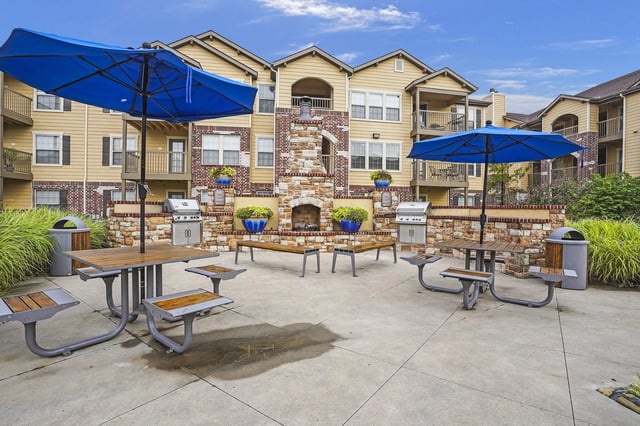 Outdoor seating area with picnic tables, umbrellas, grills, and a stone fireplace.