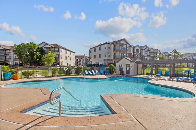 Resort-style swimming pool with lounge chairs and apartment buildings in the background.