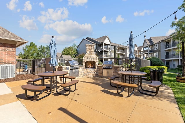 Outdoor community grilling area with picnic tables, grills, and a stone fireplace.