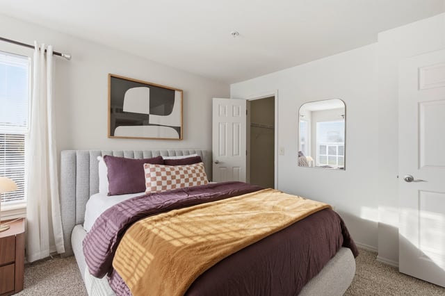 A stylish bedroom featuring a plush gray headboard, deep purple bedding with a mustard yellow throw, and modern art above the bed.