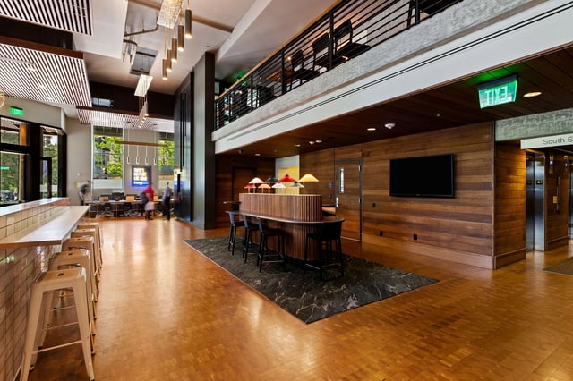 Modern lobby with wood accents, high ceilings, a seating area with bar stools, and an elevator bank.
