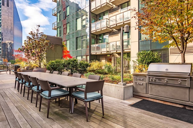 Outdoor dining area with a long table, chairs, and a grill on a wooden deck, with a modern building in the background.